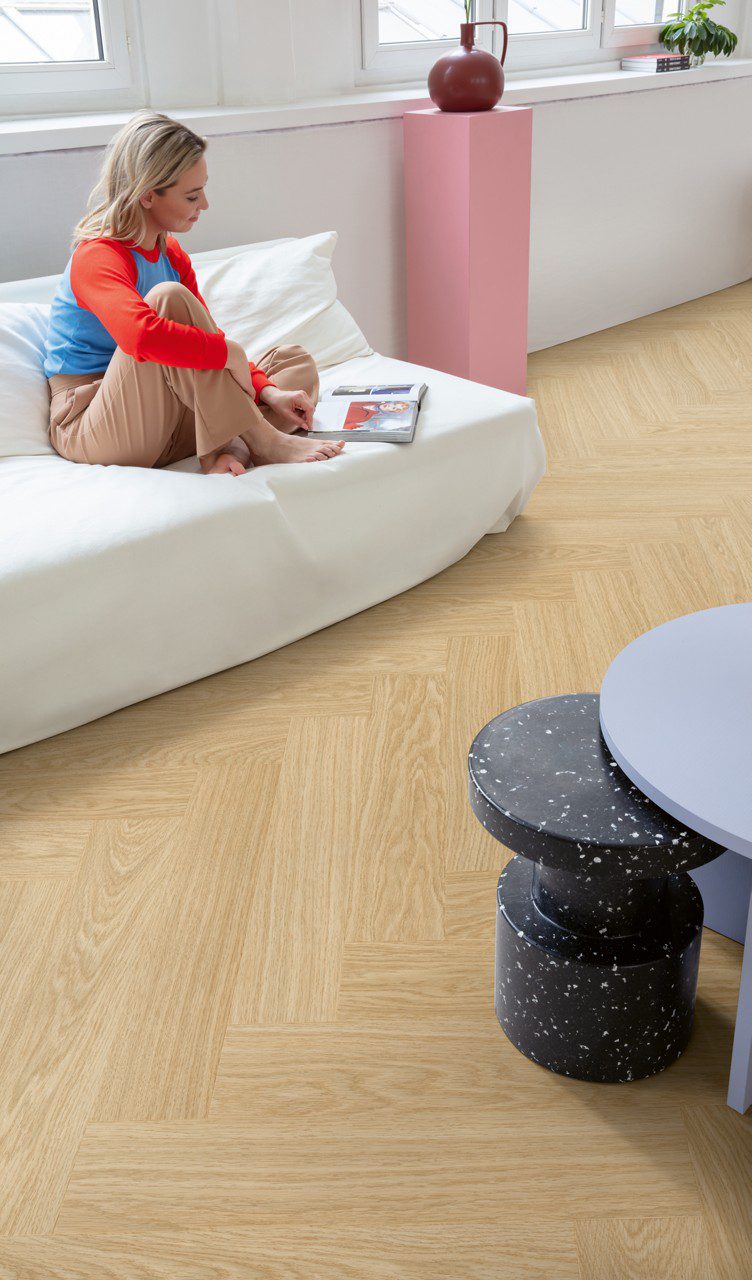 Herringbone vinyl floors Quick Step Ciro Pure Oak Blush, installed on the living room floor, a woman sitting on a white sofa browsing a magazine, a table, and a pink pedestal.