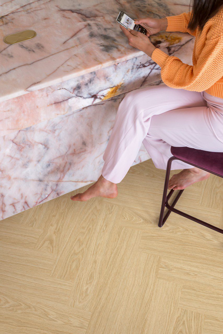 Vinyylilankku kalanruoto Quick Step Ciro Pure Oak Blush, asennettu toimiston lattialle, vaaleanpunaisesta marmorista pöytä ja paljain jaloin tuolilla istuva nainen.