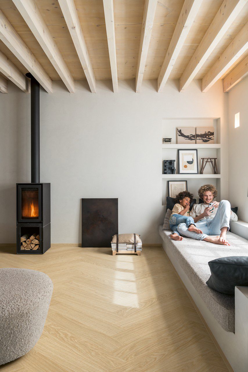 Vinyylilankku Quick Step Ciro Pure Oak Blush, asennettu olohuoneen lattialle, valurautatakassa tuli, isä ja poika makaavat sohvalla ja katsovat yhdessä puhelinta, molemmat hymyilevät.