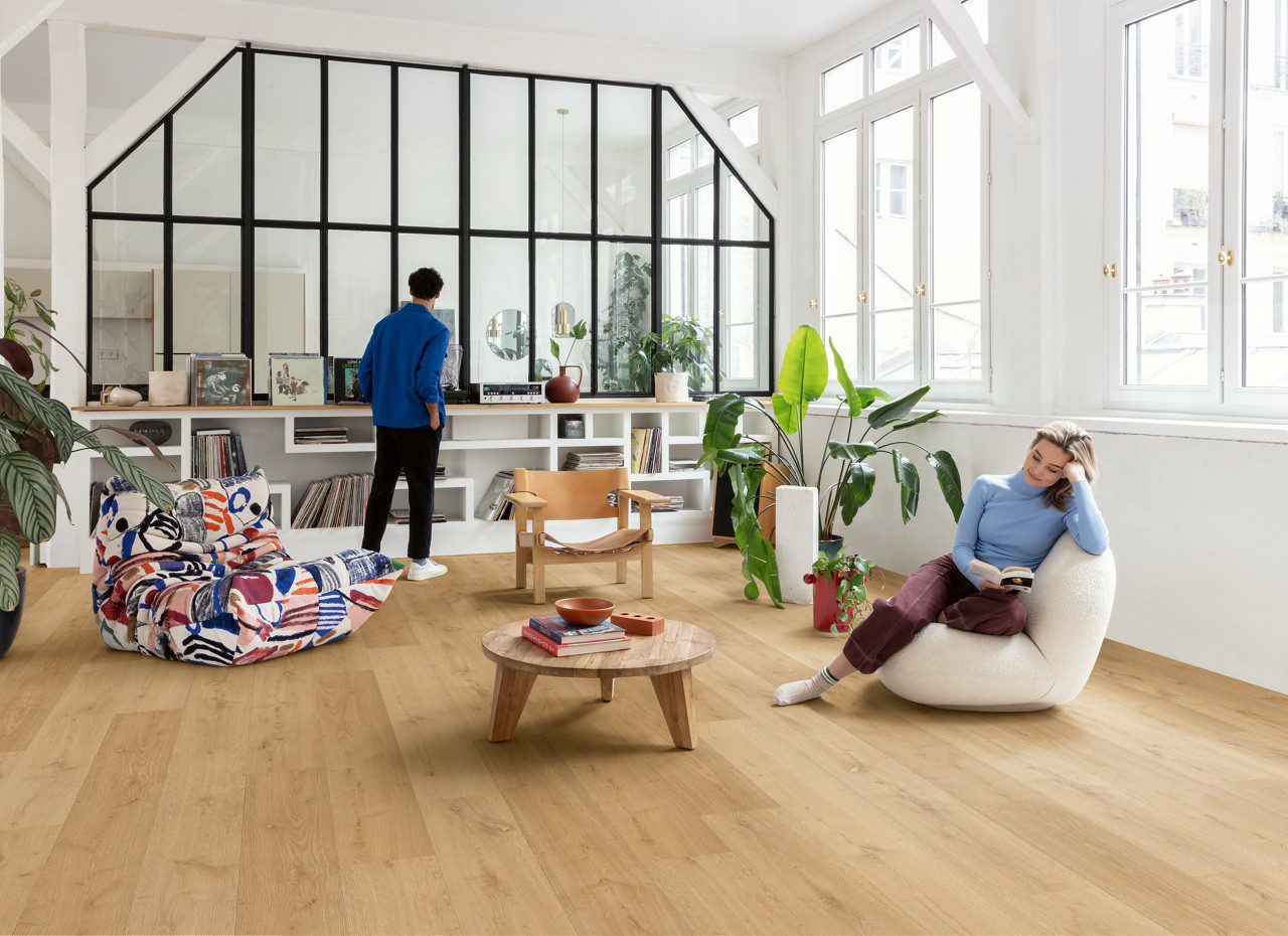 LVT floors Quick-Step Bloom Elegant Oak Natural installed on a living room floor. Woman is sitting on a chair and reading a book. Man is looking the vinyl record player. Table with books, palm threes