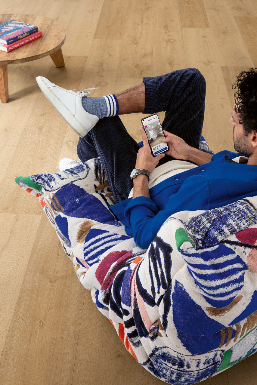 Rigid Vinyl flooring Quick-Step Bloom Elegant Oak Natural installed on ling room floor, man sitting on a chair and looking phone.