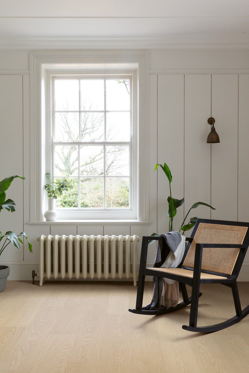 Quick Step Blos Buttermilk Oak vinyl floor installed in a living room, with a classic wall radiator, a rocking chair, white classic wall panels, and potted plants.