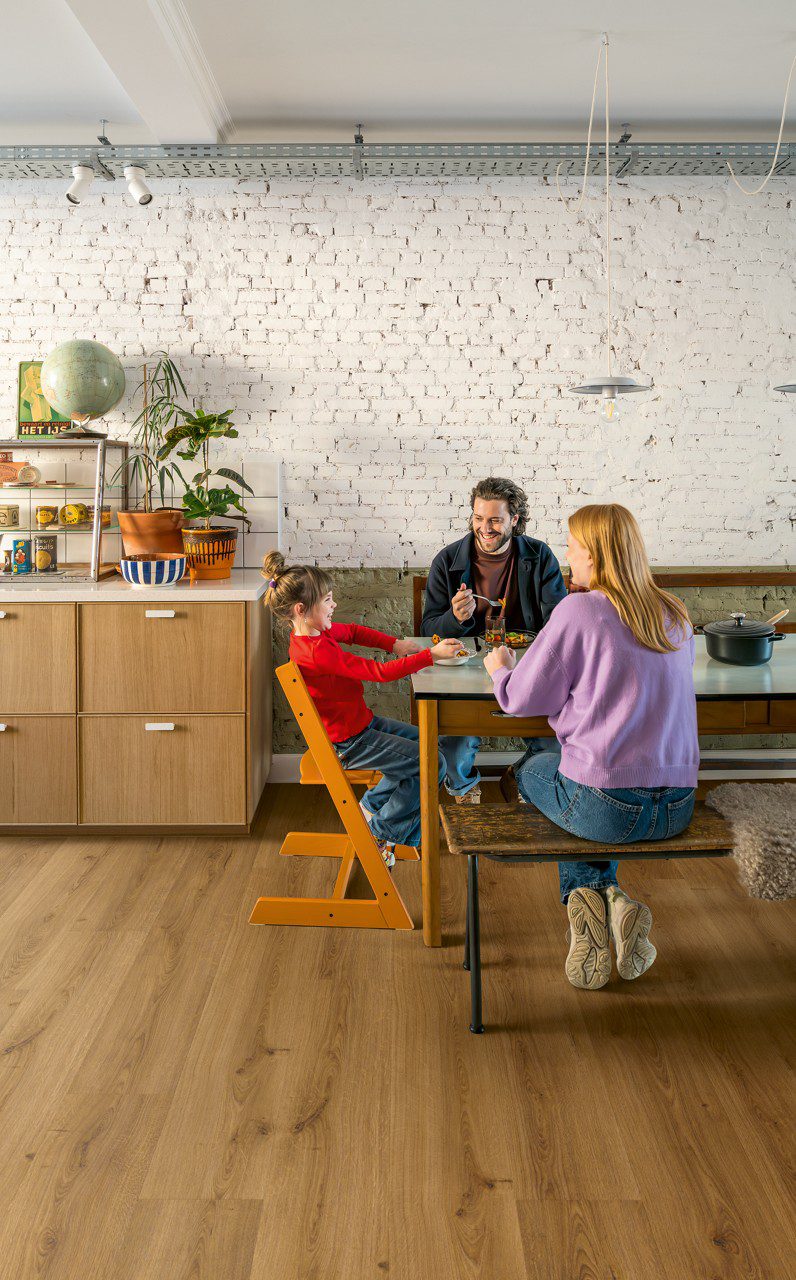 Vinyl flooring Quick-Step Bloom Botanic Caramel Oak installed on a dining room floor. Family is having dinner. Cabinets, flowers.