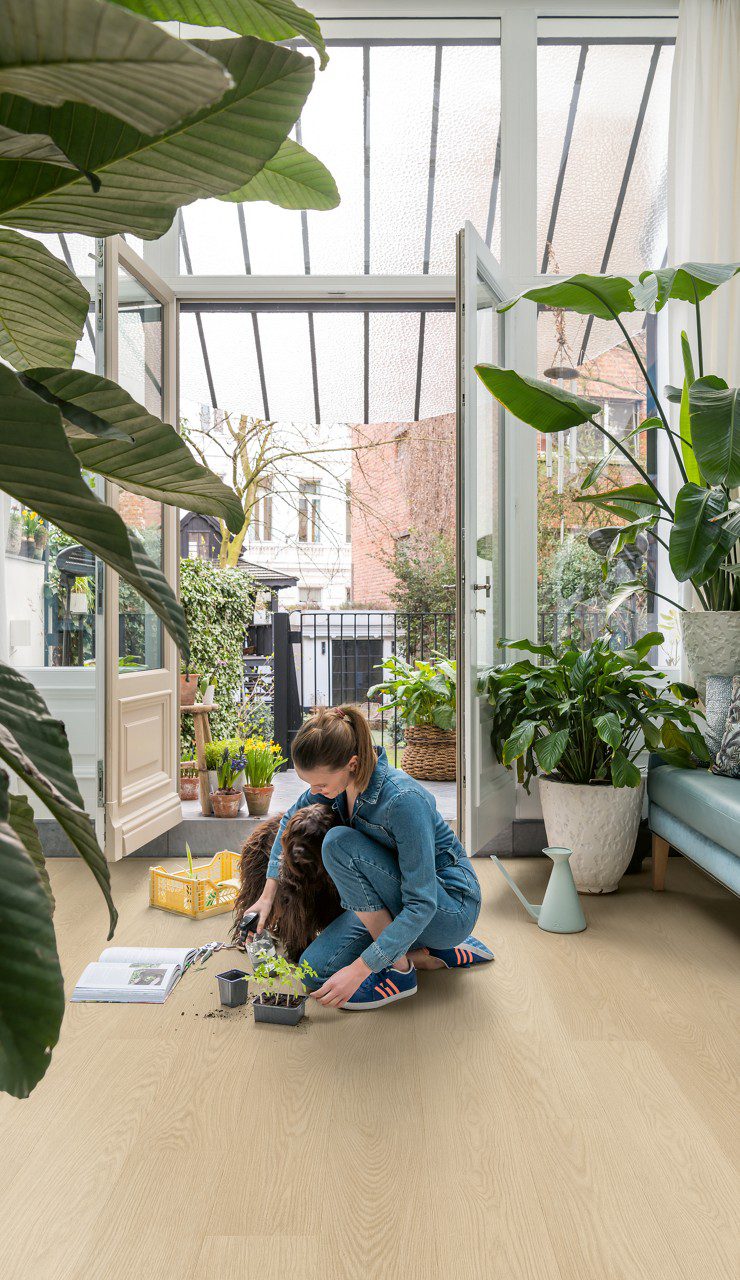 Vinyl Laminat Quick Step Blos Buttermilk Eiche im Wintergarten verlegt, viele Topfpflanzen, eine Frau pflanzt neue Pflanzen in Töpfe auf dem Boden, doppelte Balkontüren öffnen sich zum Garten mit Bäumen und Pflanzen.