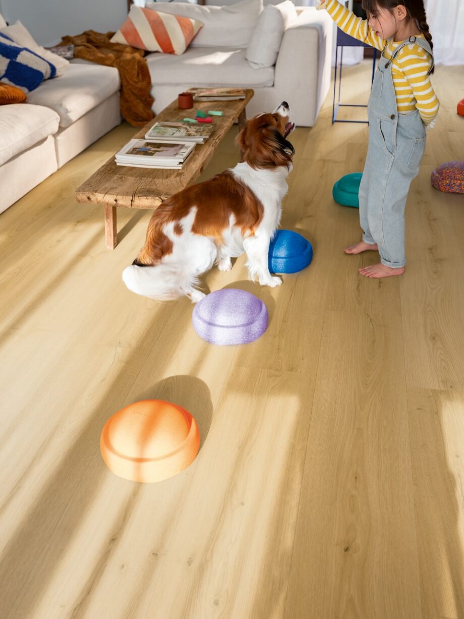 Vinylboden Quick Step Bloom Brushed Oak Honey im Wohnzimmer verlegt, Kind spielt mit einem Hund.