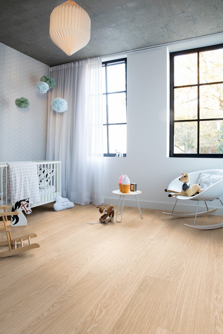 Vinyylilankku Quick Step Bloom Pure Oak Blush asennettuna lastenh uoneen lattialle, vaalea tammen sävy kevyellä punertavalla vivahteella ja lankkudesign, kehto, keinul hevonen, tuoli ja leluja.