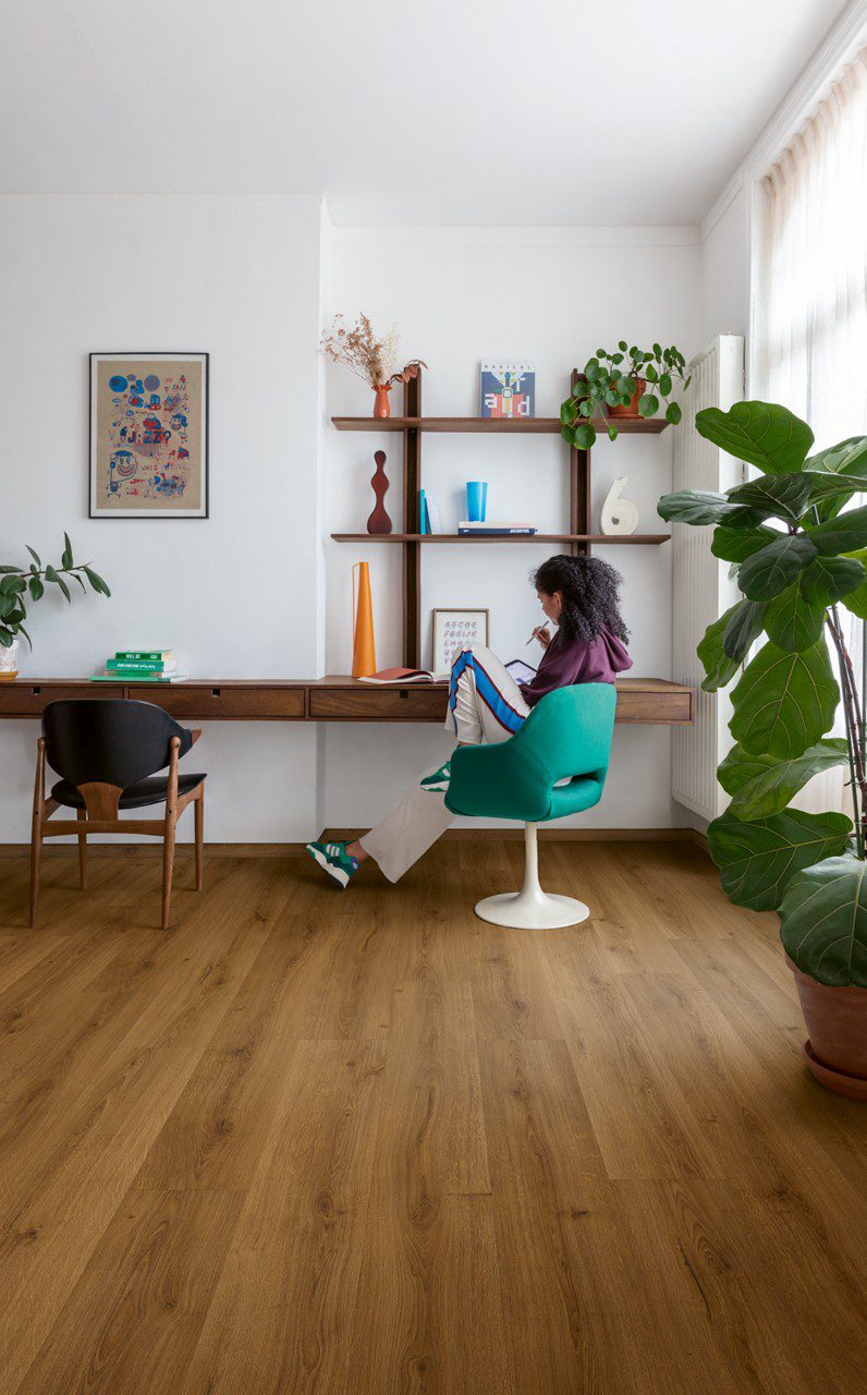 Vinyylilattia Quick Step Bloom Botanic Caramel Oak asennettuna olohuoneen lattialle, lämmin karamellinruskea tammen sävy ja lankkudesign, nainen istuu pöydän ääressä ja katsoo tablettia, palmu ja kukkia.