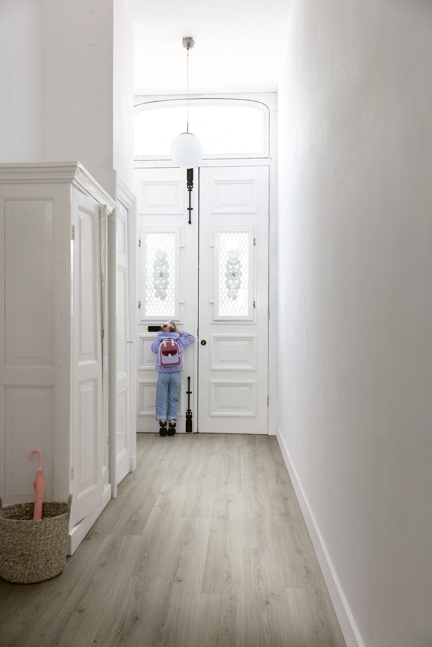 Vinyylilattia Quick Step Bloom Botanic Grey asennettuna käytävän lattialle, moderni vaaleanharmaa sävy ja lankkudesign, lapsi lähtee ulos klassisen tyylisestä ovesta.