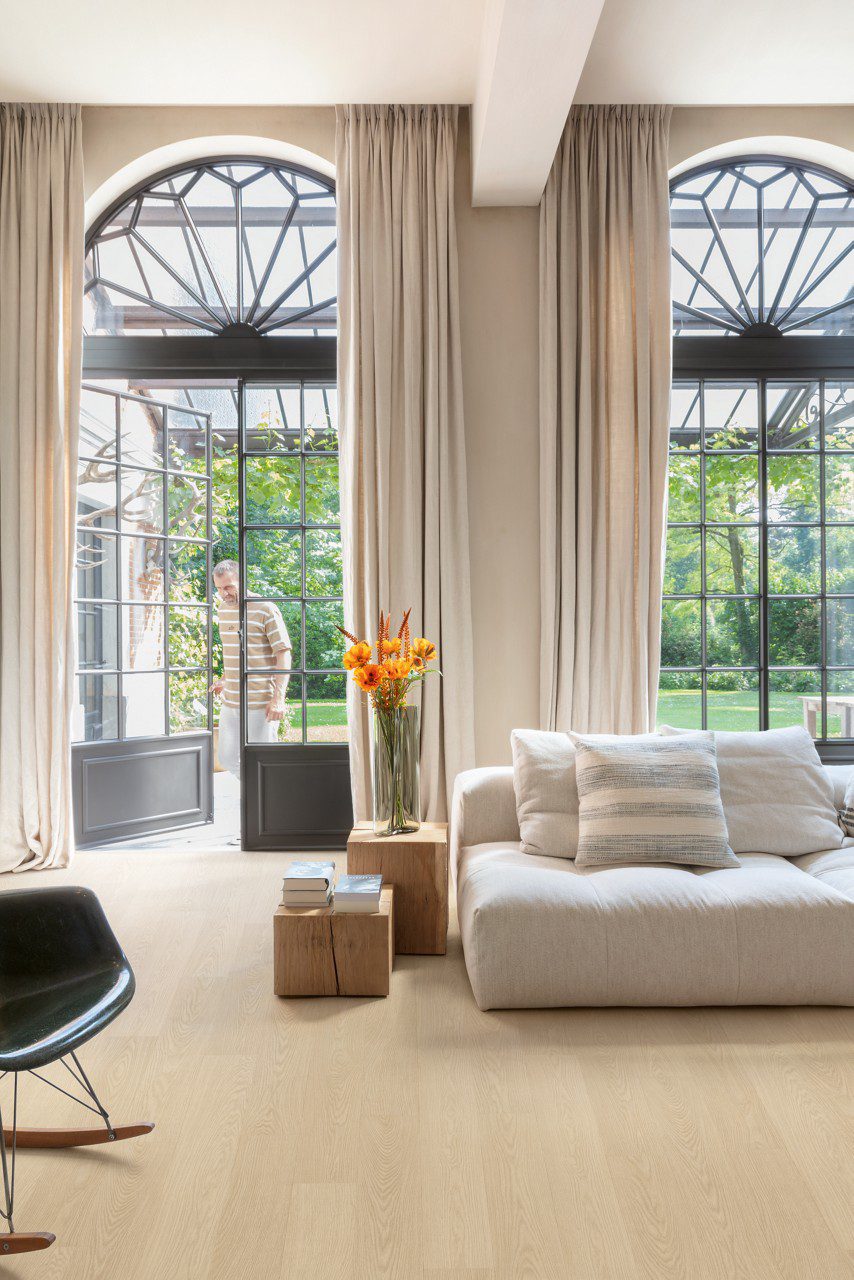 Vinyylilattia Quick Step Blos Buttermilk Vaalea Tammi asennettuna olohuoneen lattialle, vaalea kermaisen sävyinen tammen väri ja lankkudesign, kaunis sohva ja kukkia, mies astuu terassilta olohuoneeseen parvekeoven kautta.