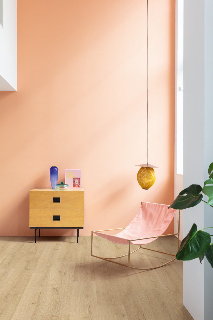 Виниловый ламинат Quick Step Bloom Botanic Beige уложен на полу в коридоре, розовый стул, комод и лампа, женщина моет пол шваброй.