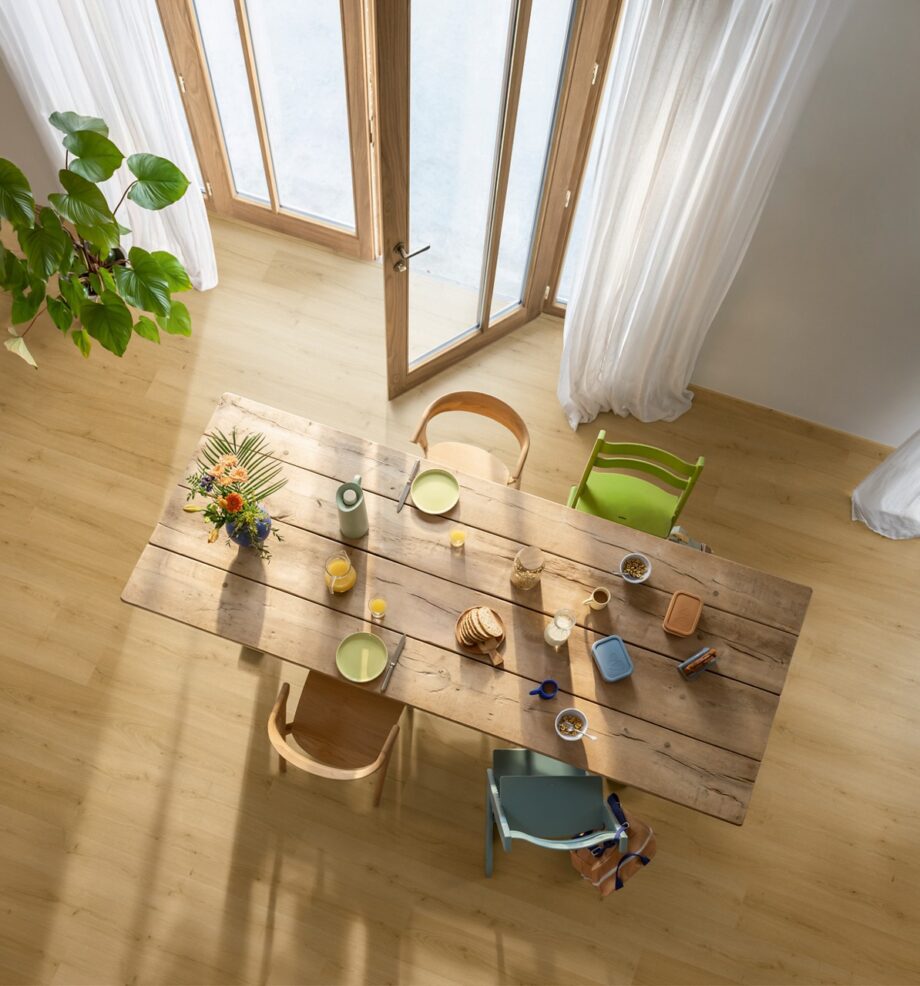 Виниловый паркет Quick Step Bloom Brushed Oak Honey уложен на полу на кухне, обеденный стол со стульями и открытая дверь на террасу.