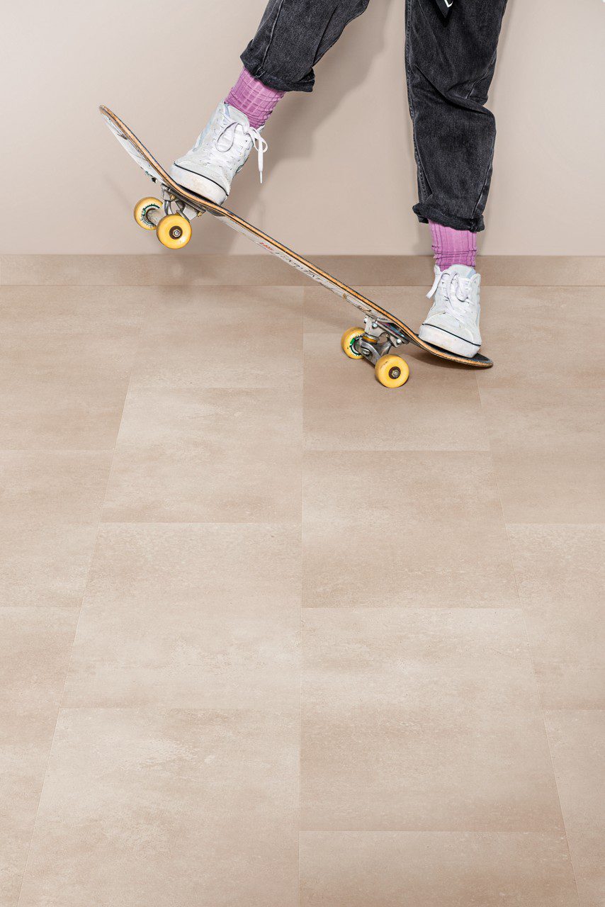 Vinylboden in Steinoptik Quick Step Oro Coral Rock auf dem Boden verlegt mit Sockelleisten im gleichen Farbton warme korall sandfarbene Steinoptik natürliche Struktur und dezente Farbvariationen Frau auf einem Skateboard