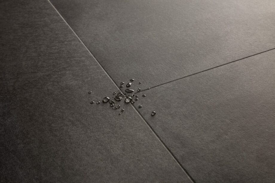 Vinyylilattia Quick-Step Illume Soft Graphite asennettuna kodin lattialle – tumma grafiitinharmaa sävy ja hillitty kivimäinen/betonimainen design hienovaraisella tekstuurilla; lattian pinnalla näkyy vesipisaroita korostamassa sen vedenkestävyyttä.