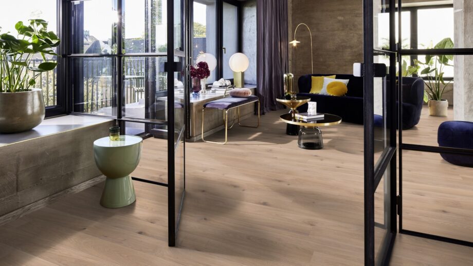 Tammeparkett Bauwerk Casapark tamm 46 Crema ültra matt lakitud, harjatud paigaldatud modernse elutoa põrandale. Lilla diivan, tumba ja pink. Kuldse äärega diivanilaud.