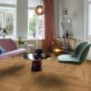 Helles, stilvolles Wohnzimmer mit Fischgrätparkett aus Eiche Mix (140 mm, geölt). Die Einrichtung kombiniert zartrosa Sofa, salbeigrünen Disainstuhl und einen modernen Couchtisch mit goldenen Akzenten. Große weiße Flügeltüren und Fenster bringen viel Tageslicht in den Raum. Warme Vorhänge, moderne Kunstobjekte und eine elegante Ambienteleuchte schaffen eine harmonische und einladende Atmosphäre. Kalasaba parkett Tamm Mix õlivahatatud 140mm – vaade 9