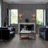 lvt-parkett-quick-step-blos-silk-tamm-dark-grey LVT parkett Quick-Step Blos Silk tamm dark grey paigaldatud klassikalise disainiga elutoa põrandale. Kaks diivanit suure diivanilaua ümber millel lilled ja küünland. Klassikalises stiilis valgest kivist kamin.