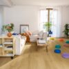 Vinyl floor Quick-Step Bloom Brushed Oak Honey installed on a living room floor. Sofa, table, flowers.
