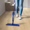 Vinylboden Quick Step Bloom Botanic Beige im Flur verlegt, eine Frau reinigt den Boden mit einem Wischmopp.