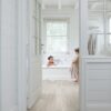 vinyylilattia-quick-step-blos-vaalea-canyon-tammi-saw-cuts Vinyylilattia Quick Step Blos Vaalea Canyon Tammi Saw Cuts asennettuna makuuhuoneen ja kylpyhuoneen lattialle, lasiovi avoinna kylpyhuoneeseen, vaahtokylpy jossa yksi lapsi leikkii ja toinen seisoo lattialla vaahtoisin jaloin.