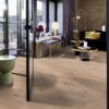 Tammeparkett Bauwerk Casapark tamm 46 Crema ültra matt lakitud, harjatud paigaldatud modernse elutoa põrandale. Lilla diivan, tumba ja pink. Kuldse äärega diivanilaud.